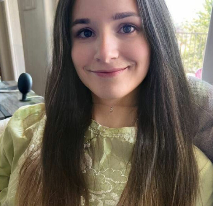 Trans Muslim woman with long dark hair and a green top smiling indoors near a window with greenery outside. Trans Muslim woman with long dark hair and a green top smiling indoors near a window with greenery outside.