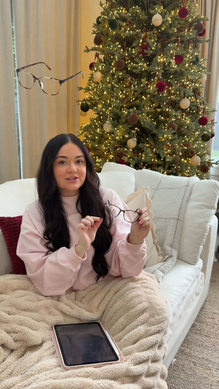 Woman on cozy sofa with Christmas tree behind, holding glasses as a recommended Christmas gift idea.