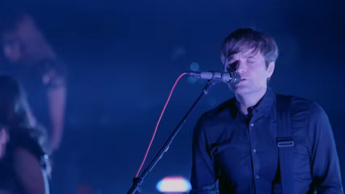 Musician singing and playing guitar on stage, unrelated to postal worker realities or delivery job challenges.