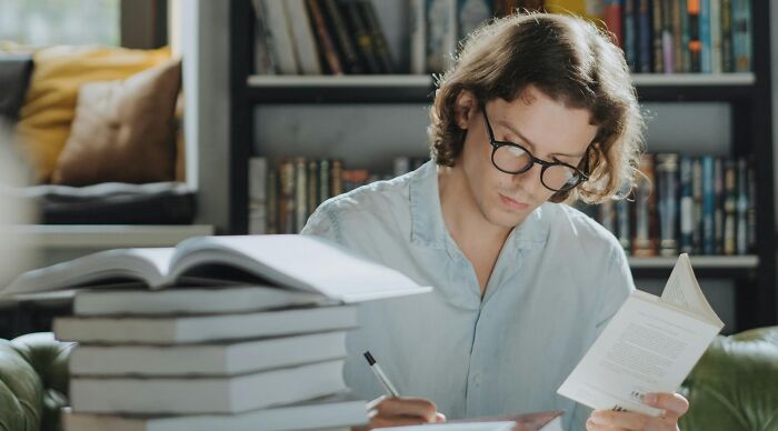 Young man with glasses solving mind-benders from a cognitive ability test, surrounded by books in a cozy study setting. Young man with glasses solving mind-benders from a cognitive ability test, surrounded by books in a cozy study setting.