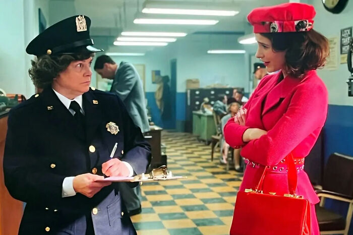 Actress from Marvelous Mrs. Maisel wearing red outfit standing in a police station talking to female officer with clipboard. Actress from Marvelous Mrs. Maisel wearing red outfit standing in a police station talking to female officer with clipboard.