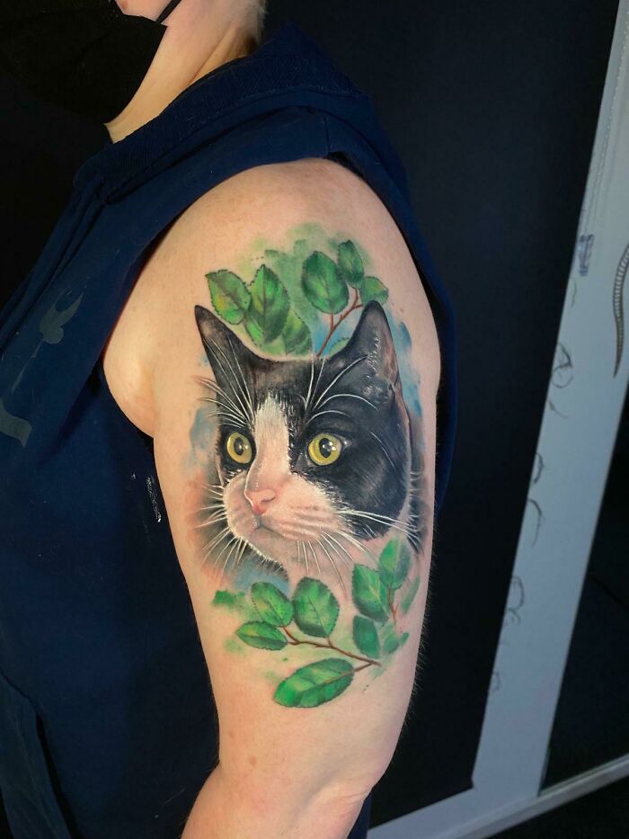 Realistic tattoo of a black and white cat surrounded by green leaves, a beautiful tribute to a beloved pet memory on arm.
