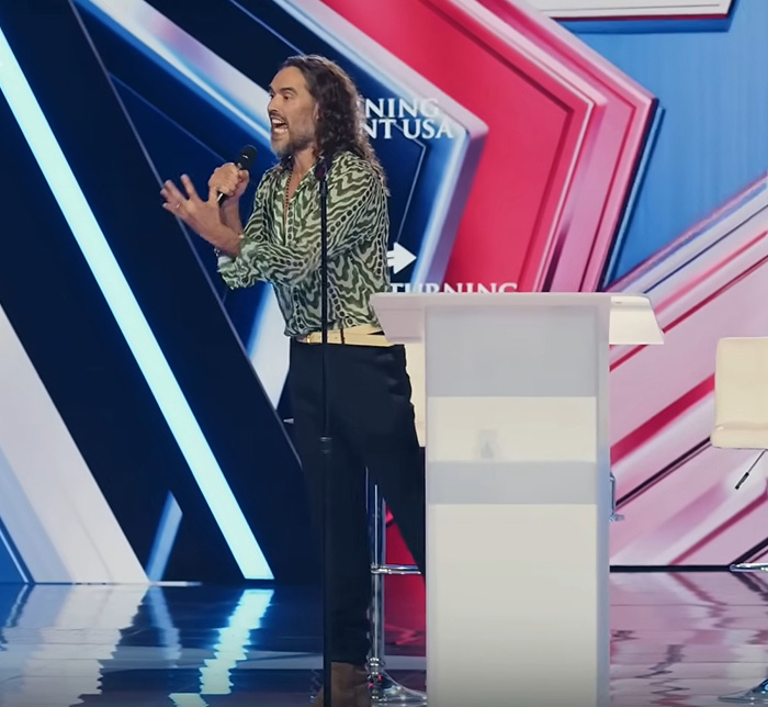 Russell Brand speaking passionately on stage, reacting to ex-wife Katy Perry's new relationship with Justin Trudeau.