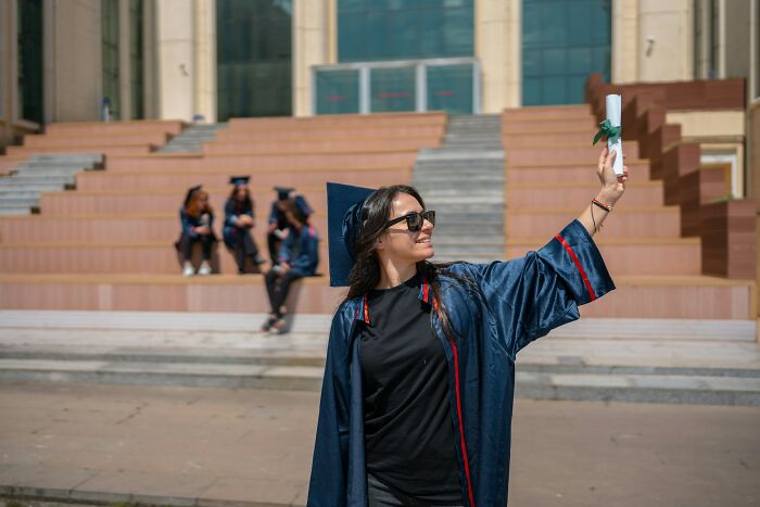 Graduate woman in cap and gown holding diploma outdoors with peers in the background in a true stories that sound made up setting.