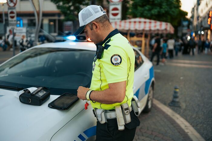 Police officer wearing bright yellow uniform standing next to patrol car using digital tablet representing instant karma concept.