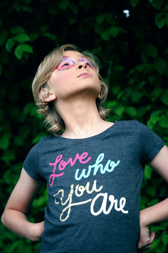 Child with colorful face paint and bold pose, wearing a love who you are shirt amid green leafy background, reflecting transgender baby theme.