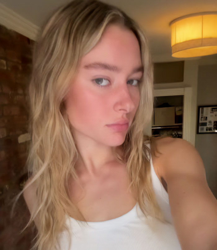 Young woman with long blonde hair wearing a white tank top inside a dimly lit room, related to NYU shove victim friend. Young woman with long blonde hair wearing a white tank top inside a dimly lit room, related to NYU shove victim friend.