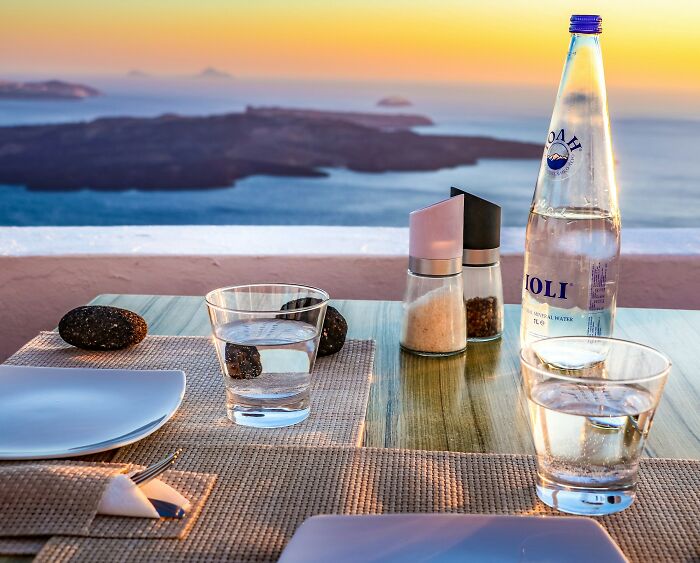 Table set with water glasses, salt, pepper, and bottle overlooking sea at sunset in a Greek village setting. Table set with water glasses, salt, pepper, and bottle overlooking sea at sunset in a Greek village setting.