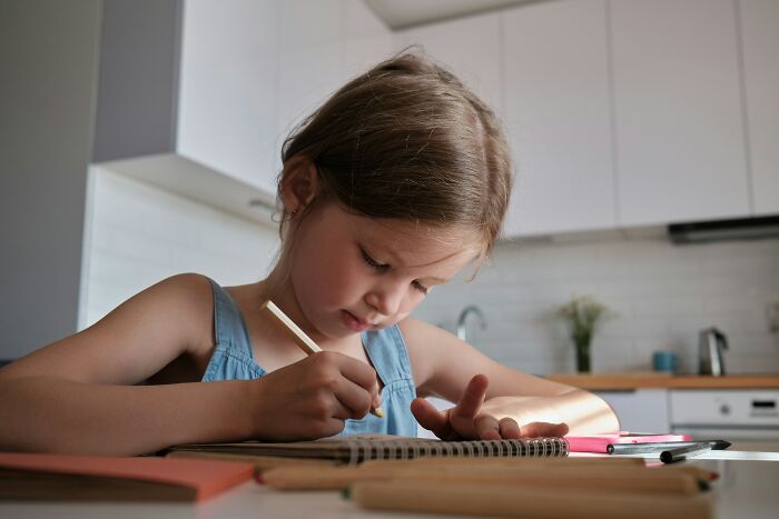 Young girl focused on writing a strange and specific kid's insult in her notebook at a bright kitchen table.