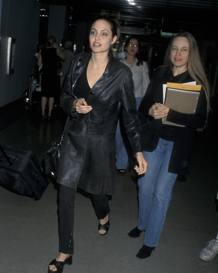 Angelina Jolie walking in a black leather coat with a serious expression, highlighting strength after revealing mastectomy scars.