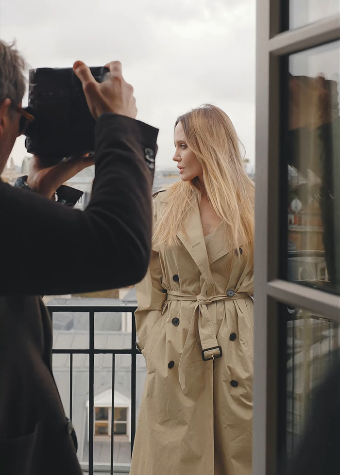 Angelina Jolie in a beige trench coat posing outdoors while a photographer captures images on a balcony.
