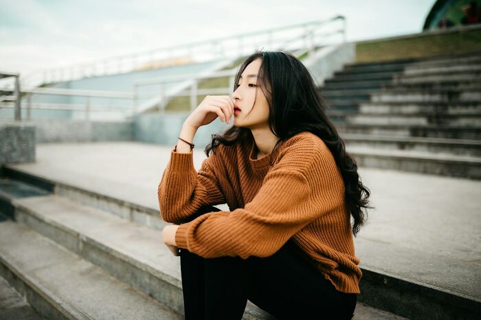 Young woman in a brown sweater sitting on concrete steps, lost in thought, reflecting on unsolved mysteries and puzzles.