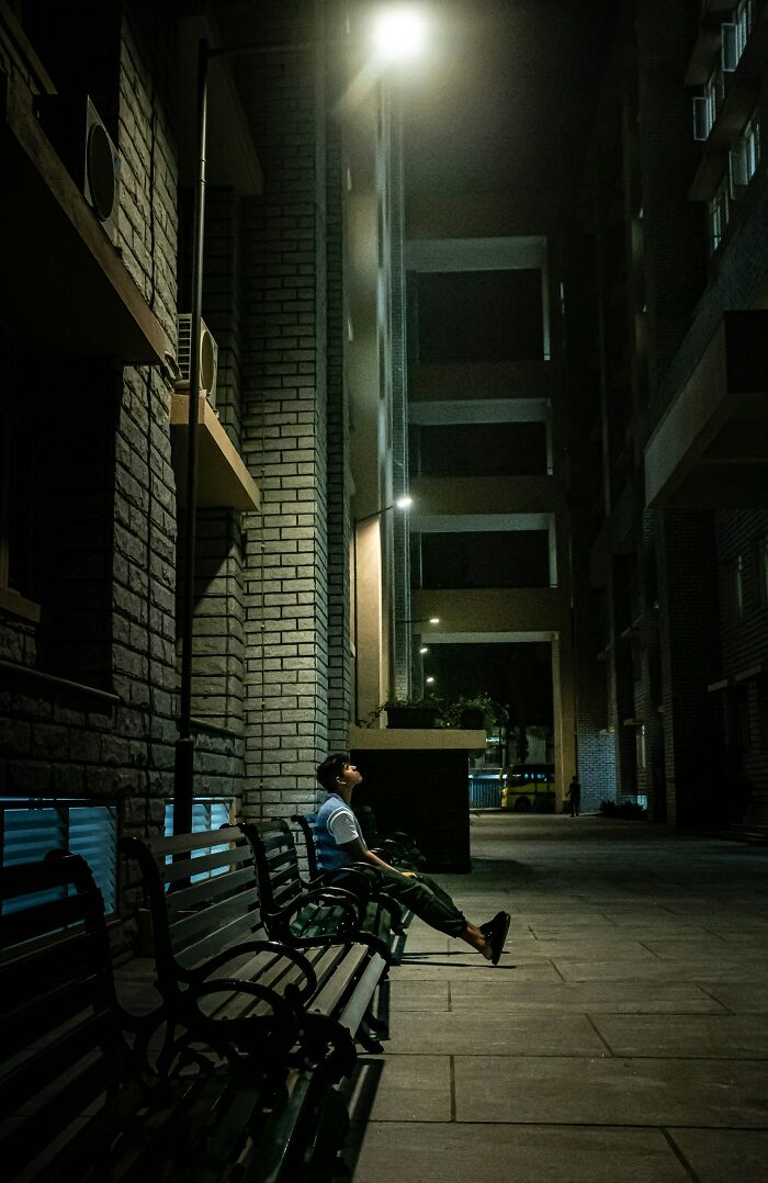 Person sitting alone on a bench at night between tall buildings, reflecting on backstabbing and regretful actions.