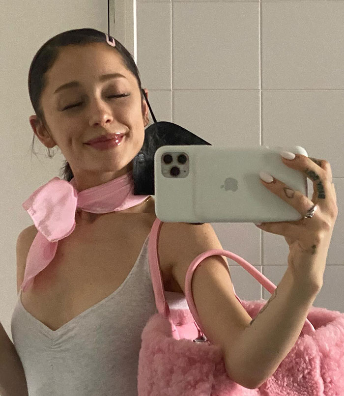 Ariana Grande taking a mirror selfie holding a phone, wearing a pink scarf and carrying a fluffy pink bag against a tiled background.