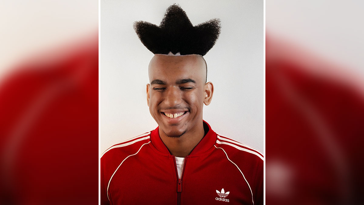 Young man with bold barber haircut styled into a star shape wearing a red Adidas jacket smiling with eyes closed.
