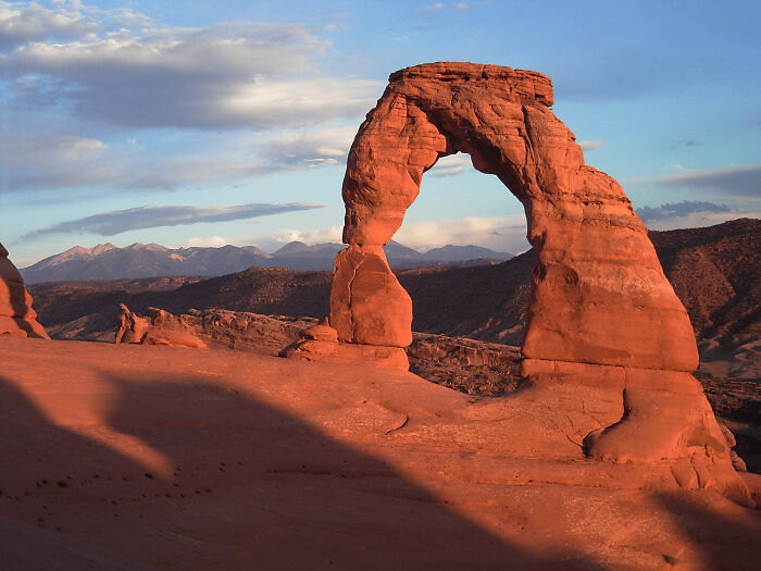 Natural red rock arch formation under a partly cloudy sky, one of the places that look AI generated but are actually real.