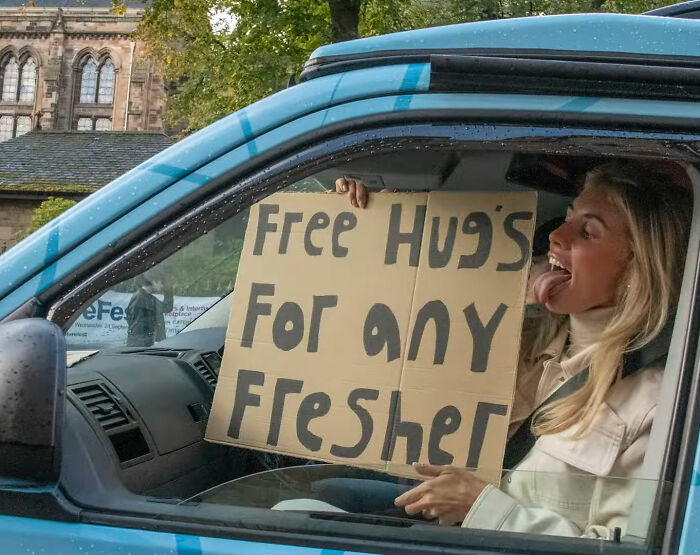 Woman sitting in a blue car holding a cardboard sign offering free hugs, unrelated to Bonnie Blue adult video Bali sentence news.