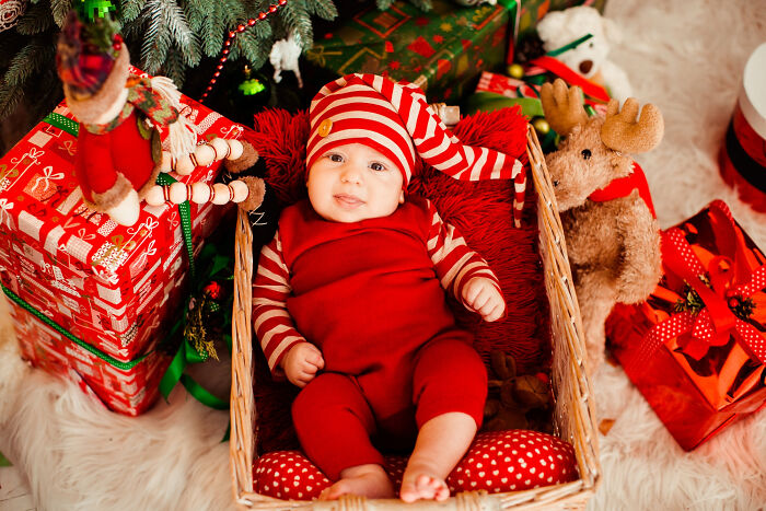 Baby dressed in red lying in basket under Christmas tree surrounded by holiday gifts and decorations. Baby dressed in red lying in basket under Christmas tree surrounded by holiday gifts and decorations.