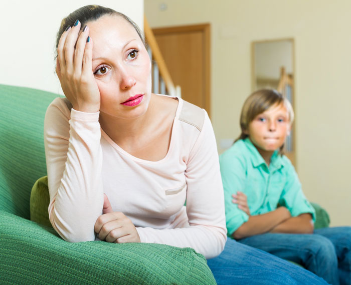 Woman looking stressed sitting on a couch with a boy in the background, illustrating family expecting free babysitting. Woman looking stressed sitting on a couch with a boy in the background, illustrating family expecting free babysitting.