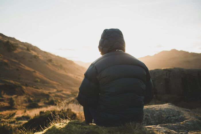 Person wearing a black jacket sitting on a rock, overlooking a mountainous landscape at sunset, reflecting on bizarre names.