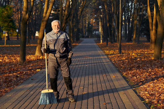 Elderly man standing on a park path holding a broom, surrounded by autumn leaves, showing resilience and calm.