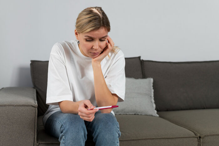 Woman sitting on couch looking worried while holding a pregnancy test, reflecting on biggest bullet dodged moments.