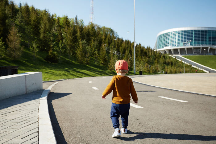 Young child walking alone on a curved road near modern building and green trees, symbolizing dodging a big bullet unnoticed.