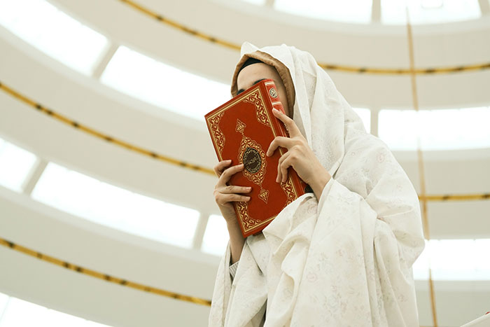 Person in white cloak holding a red ornate book close to their face inside a circular building interior.