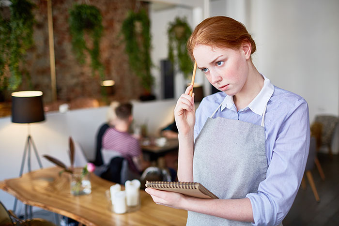Young waitress wearing apron thoughtfully holding pencil and notepad in restaurant with customers dining in background