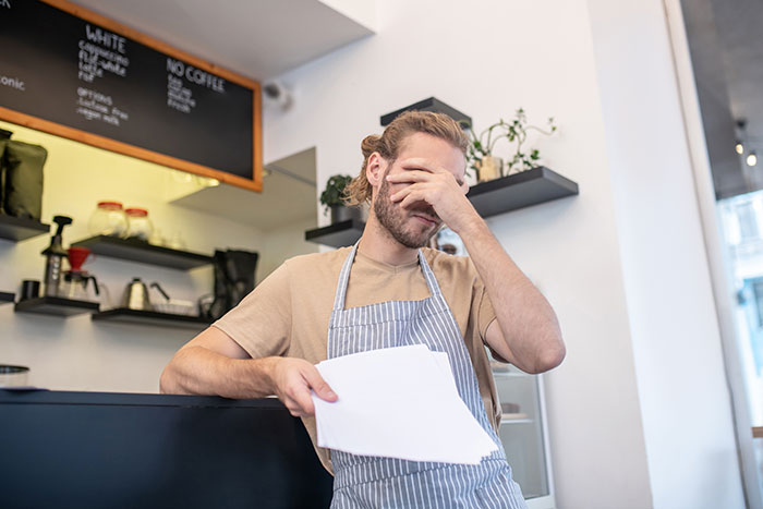 Young male waitress in apron covering face in frustration inside a cafe after entitled customer forgets manners.