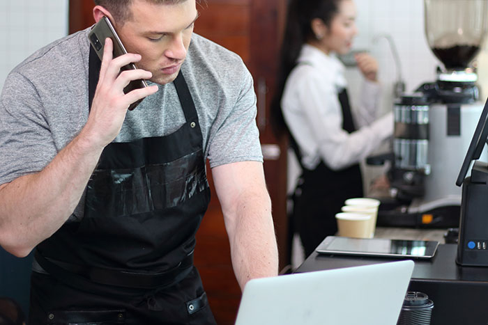 Barista wearing black apron talking on phone while looking at laptop, serving staff handling orders in background Barista wearing black apron talking on phone while looking at laptop, serving staff handling orders in background