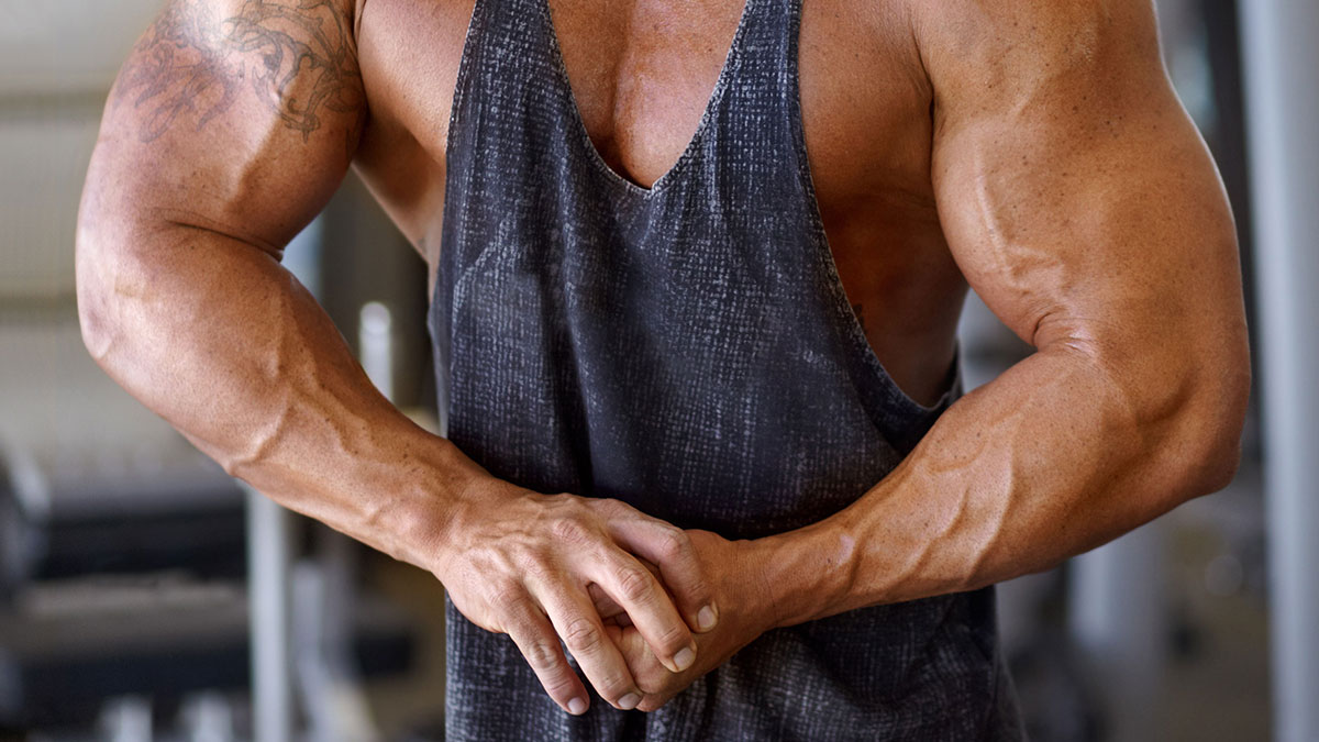 Muscular man with tattooed arm wearing tank top, clenching hands in gym, reflecting tension on future childu2019s health risk.