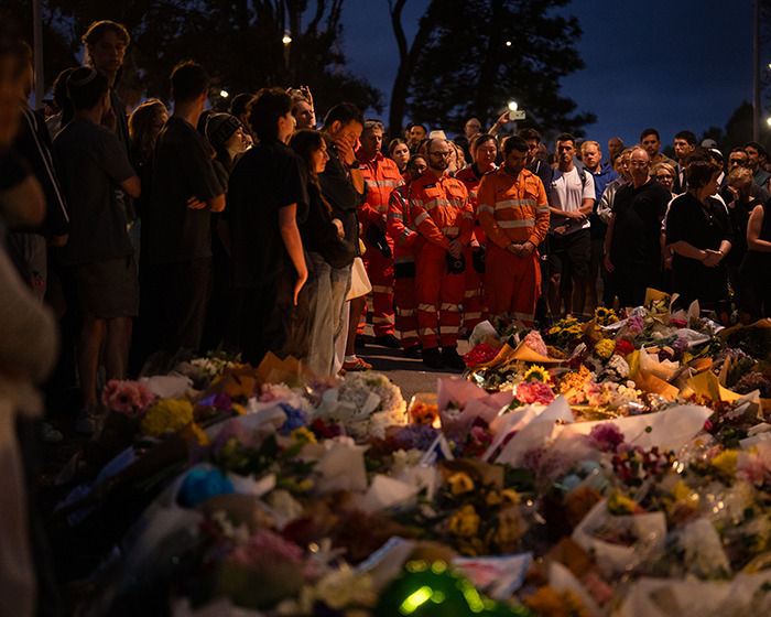 Crowd gathers at a memorial on Bondi Beach, honoring hero who confronted attacker, with emergency responders present at night. Crowd gathers at a memorial on Bondi Beach, honoring hero who confronted attacker, with emergency responders present at night.
