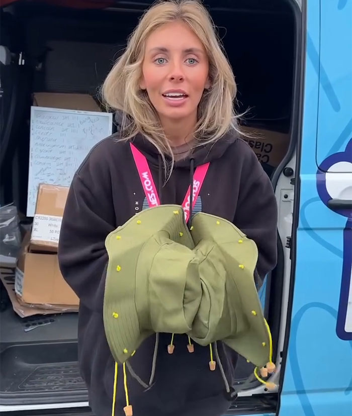 Blonde woman delivering Bonnie Blue's defiant message for authorities while holding green hats near a van in daylight. Blonde woman delivering Bonnie Blue's defiant message for authorities while holding green hats near a van in daylight.