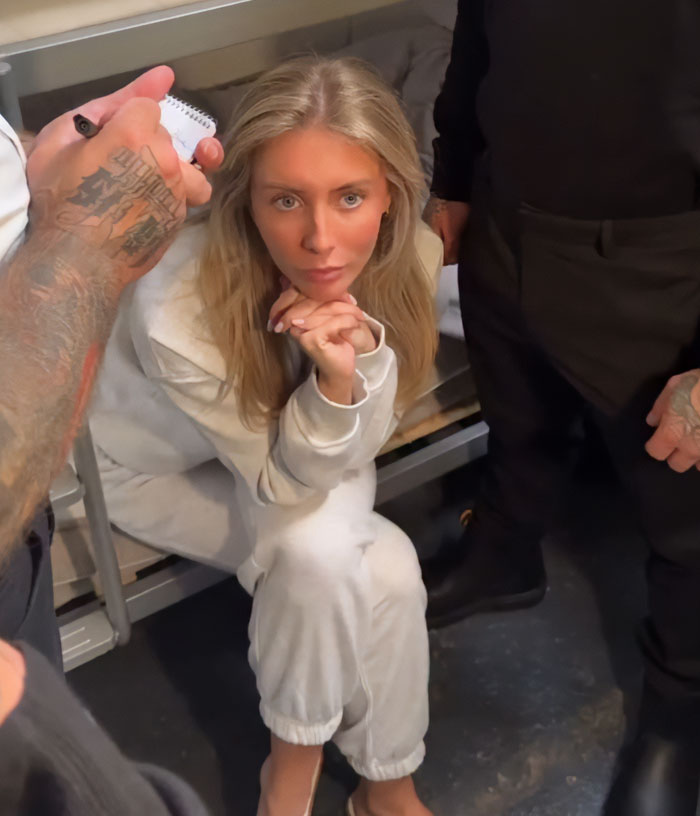 Bonnie Blue sitting in a white outfit, looking defiant while surrounded by tattooed men in a dimly lit room. Bonnie Blue sitting in a white outfit, looking defiant while surrounded by tattooed men in a dimly lit room.