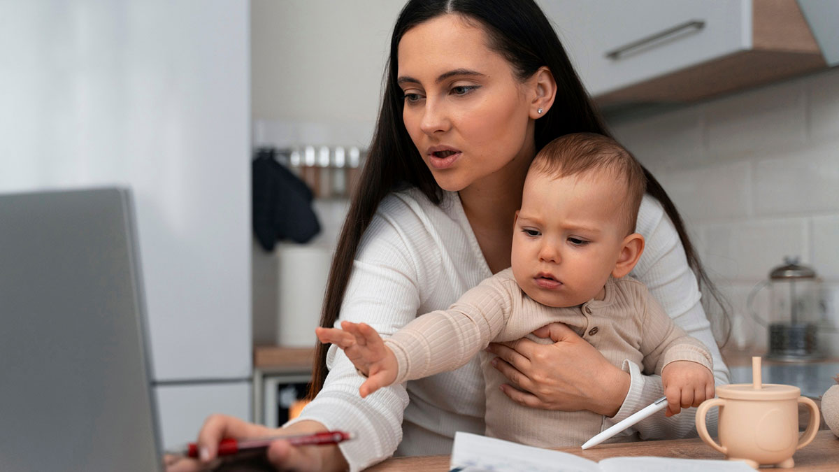 Young woman with child working on laptop at home while multitasking and attending to baby in casual setting.