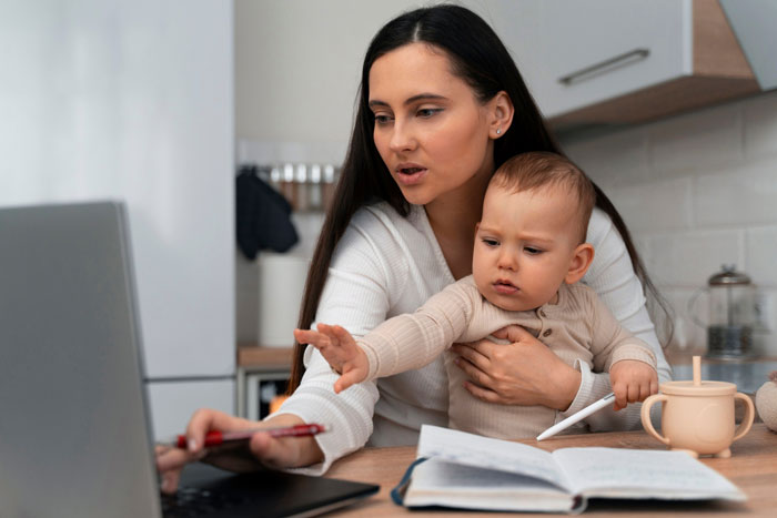Woman holding baby while working on laptop at home, relating to boss asking lady to host family for a week. Woman holding baby while working on laptop at home, relating to boss asking lady to host family for a week.