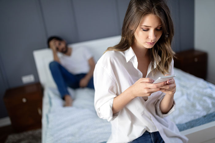 Woman upset using phone in bedroom while boyfriend sits on bed blaming her Cancer zodiac sign for cheating issues.