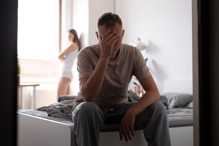 Man in tears sitting on bed covering face while female colleague stands distant, highlighting misogyny and jealousy conflict.
