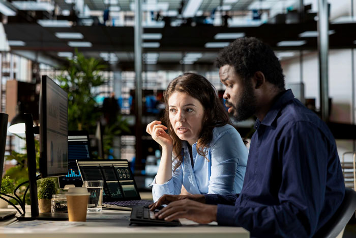A male and female colleague discussing data on computer screens in an office, highlighting misogyny workplace tension.