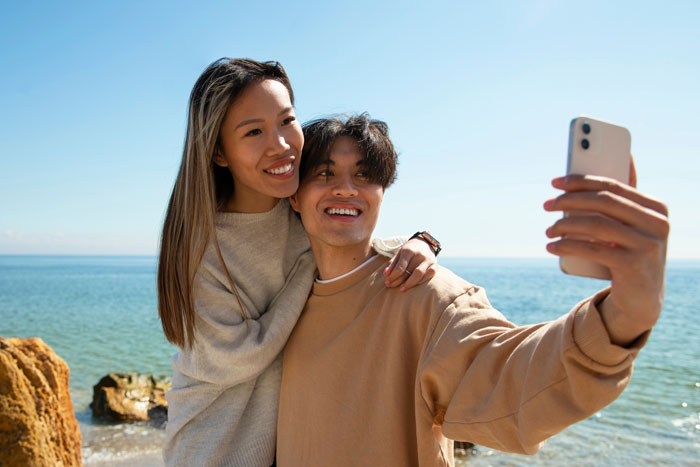 Young couple taking a selfie at the beach, boyfriend editing face every photo with a smile and affectionate pose. Young couple taking a selfie at the beach, boyfriend editing face every photo with a smile and affectionate pose.