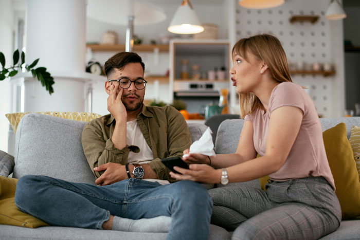 A frustrated boyfriend editing face on every photo while sitting on a couch with a woman talking to him at home. A frustrated boyfriend editing face on every photo while sitting on a couch with a woman talking to him at home.