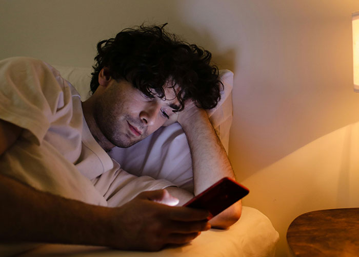 A man lying in bed at night, looking at his phone with a sad expression, reflecting on a crush at work.