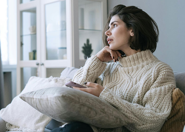 Young woman in cozy sweater holding phone, looking thoughtful while sitting on a couch at home, reflecting on relationships.