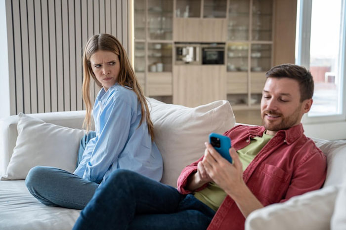 Angry girlfriend looks horrified while boyfriend scrolls phone on couch, tense moment about a racist gift. Angry girlfriend looks horrified while boyfriend scrolls phone on couch, tense moment about a racist gift.