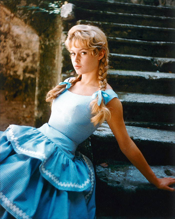Brigitte Bardot in a blue dress with braids, seated on outdoor stone stairs in a vintage style portrait.