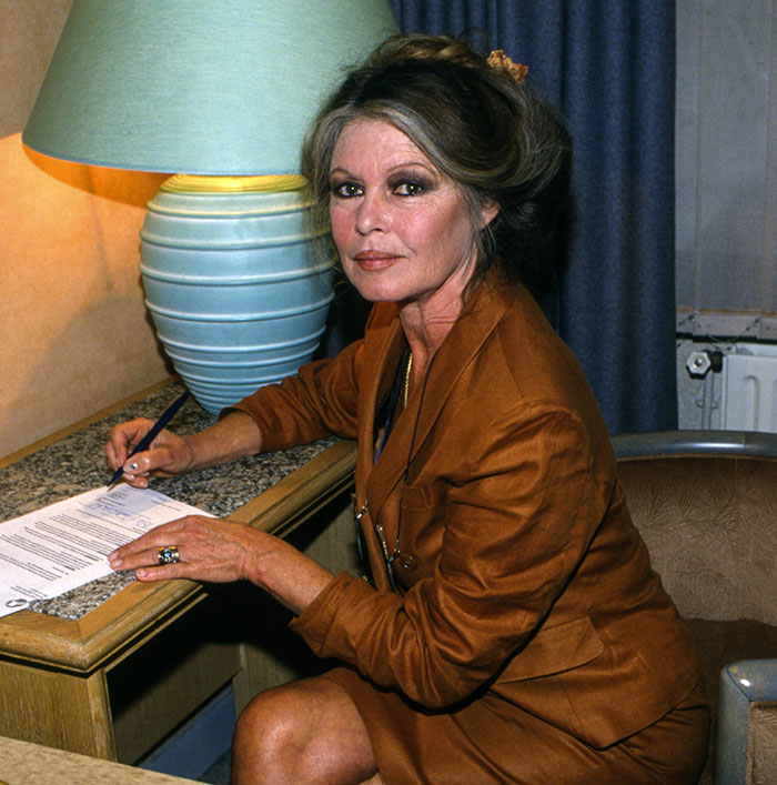 Brigitte Bardot sitting at desk signing papers with a lamp behind her, related to backlash over Chappell Roan post.