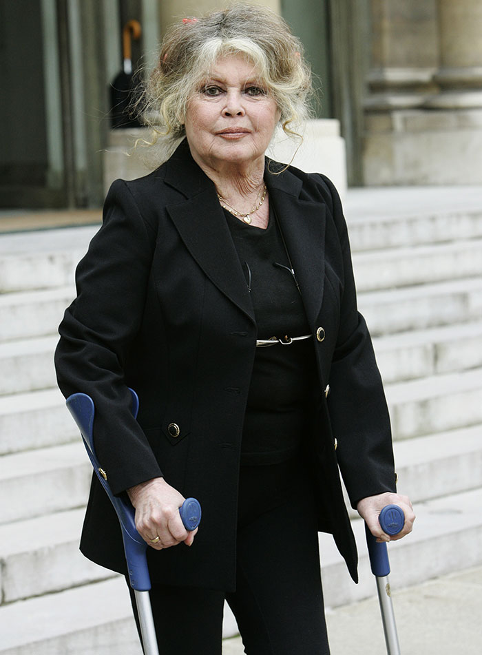 Brigitte Bardot wearing black outfit using crutches while standing on outdoor steps in daylight.
