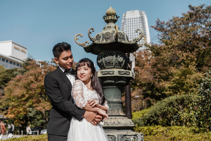 Groom embracing his wife in wedding dress by ornate lantern in sunny park, city buildings and trees in background