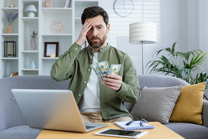 Man in casual clothes sitting at home, stressed and counting cash, reflecting financial instability and relationship pressure.